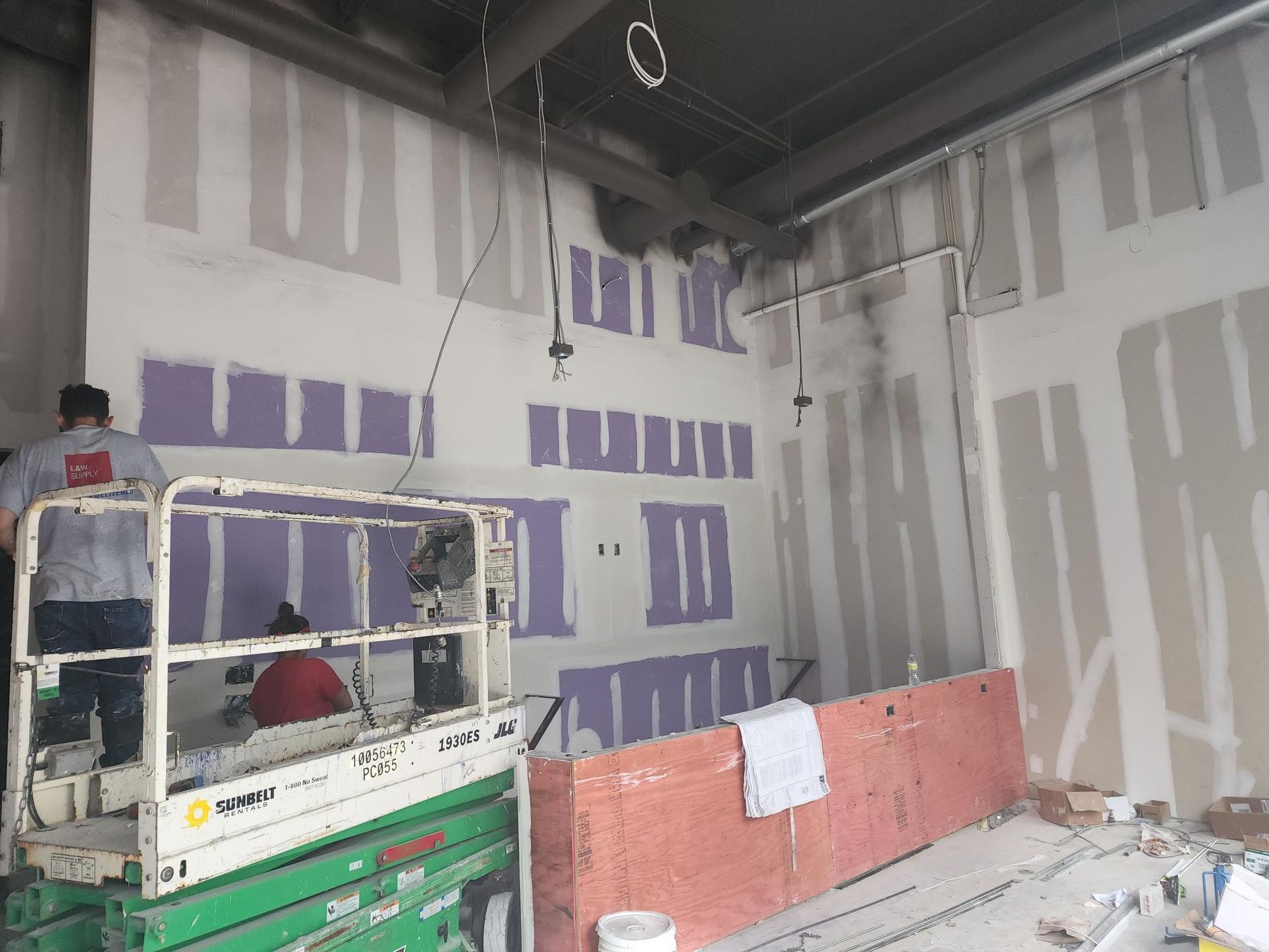 Construction site interior with drywall patches, workers on lifts, and orange safety barriers