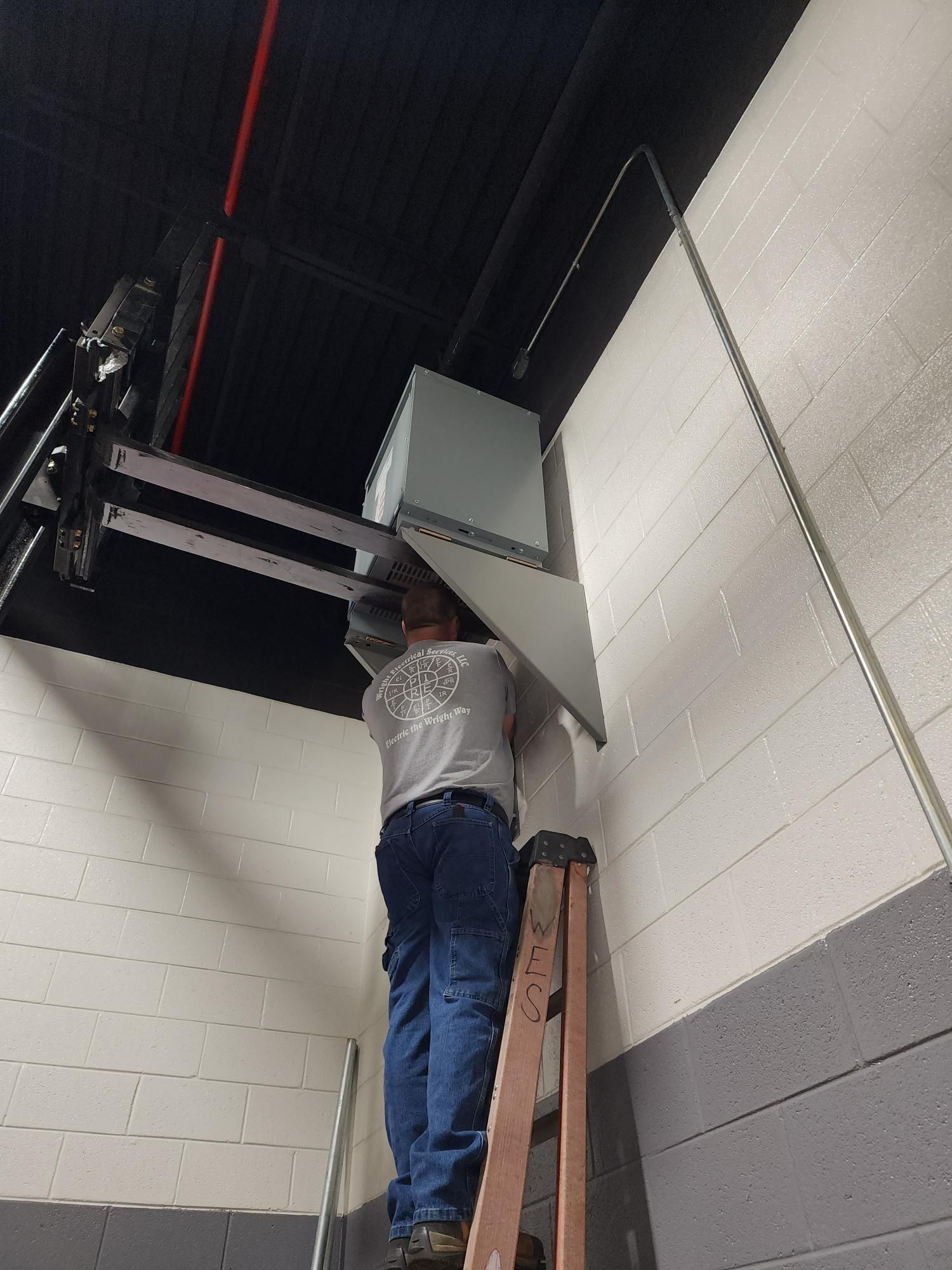 Person on a ladder installing a gray box on a high cinder block wall in an industrial room