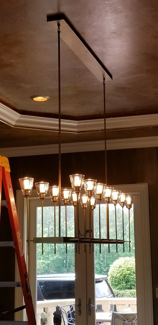 Chandelier with multiple lights hanging in a room with a window, ladder on the left, and a decorative ceiling.