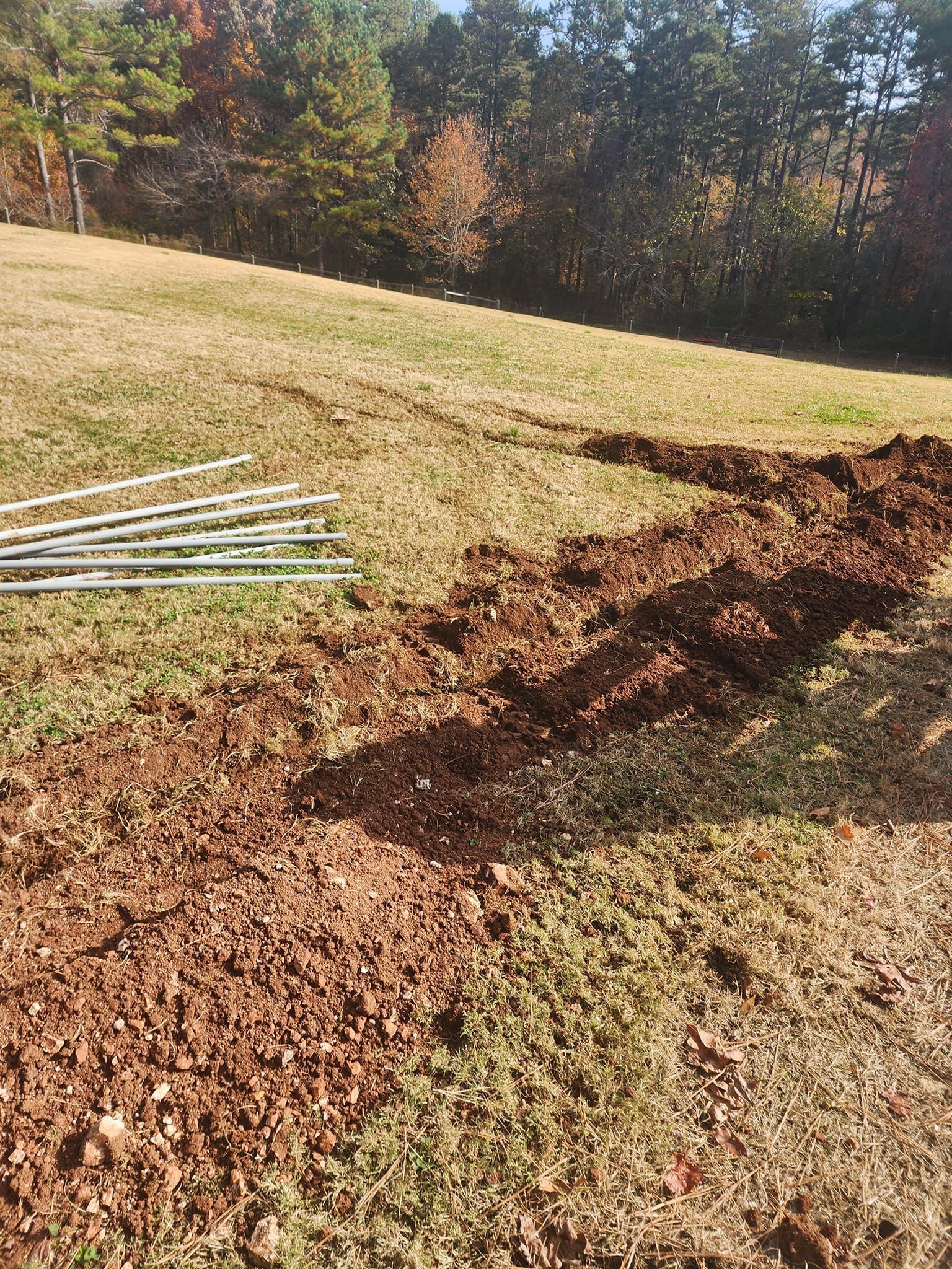 A trench dug in a grassy yard, with metal pipes on the left and trees in the background.