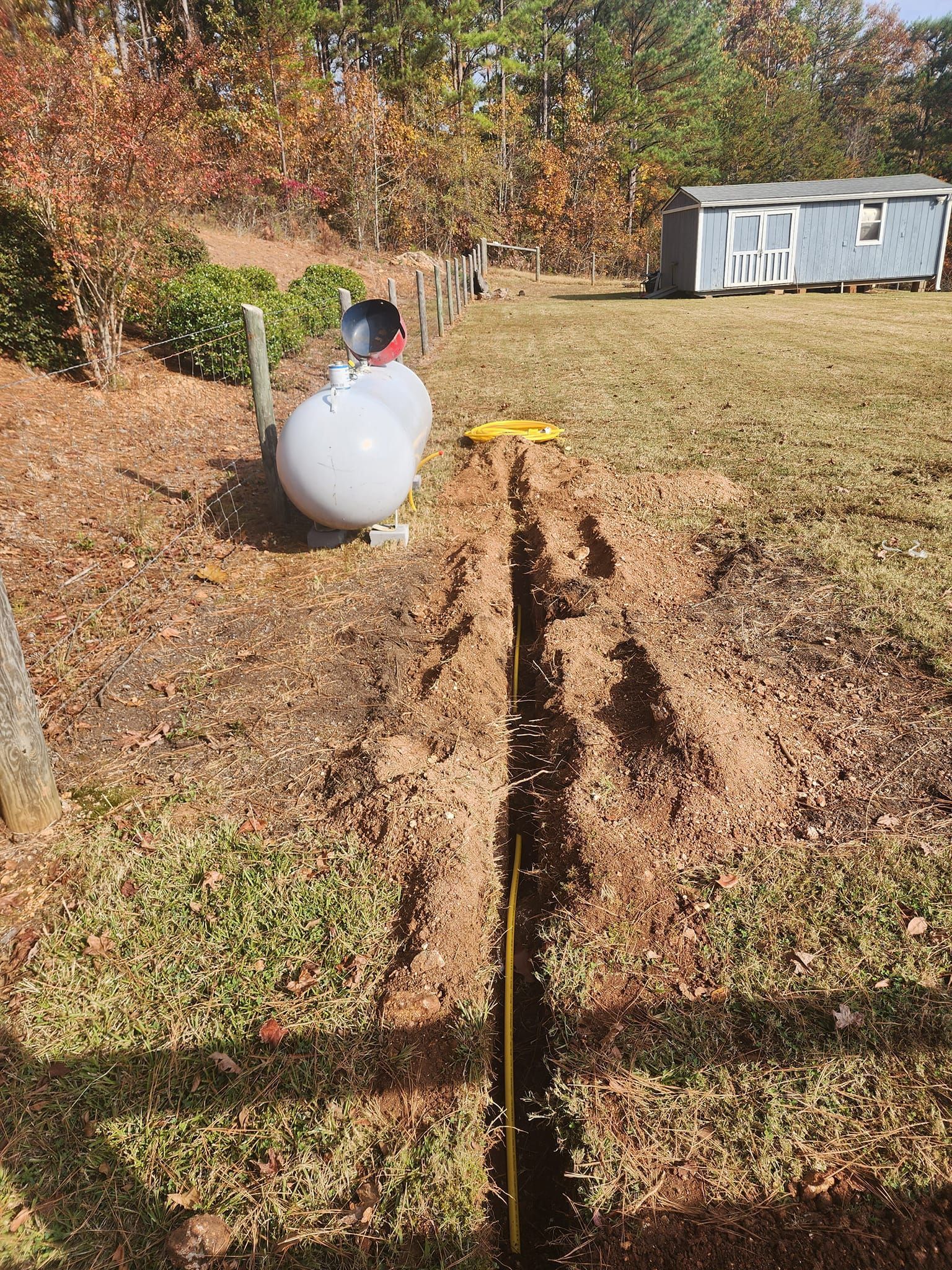 A propane tank next to a trench dug in a yard, with a small building in the background.