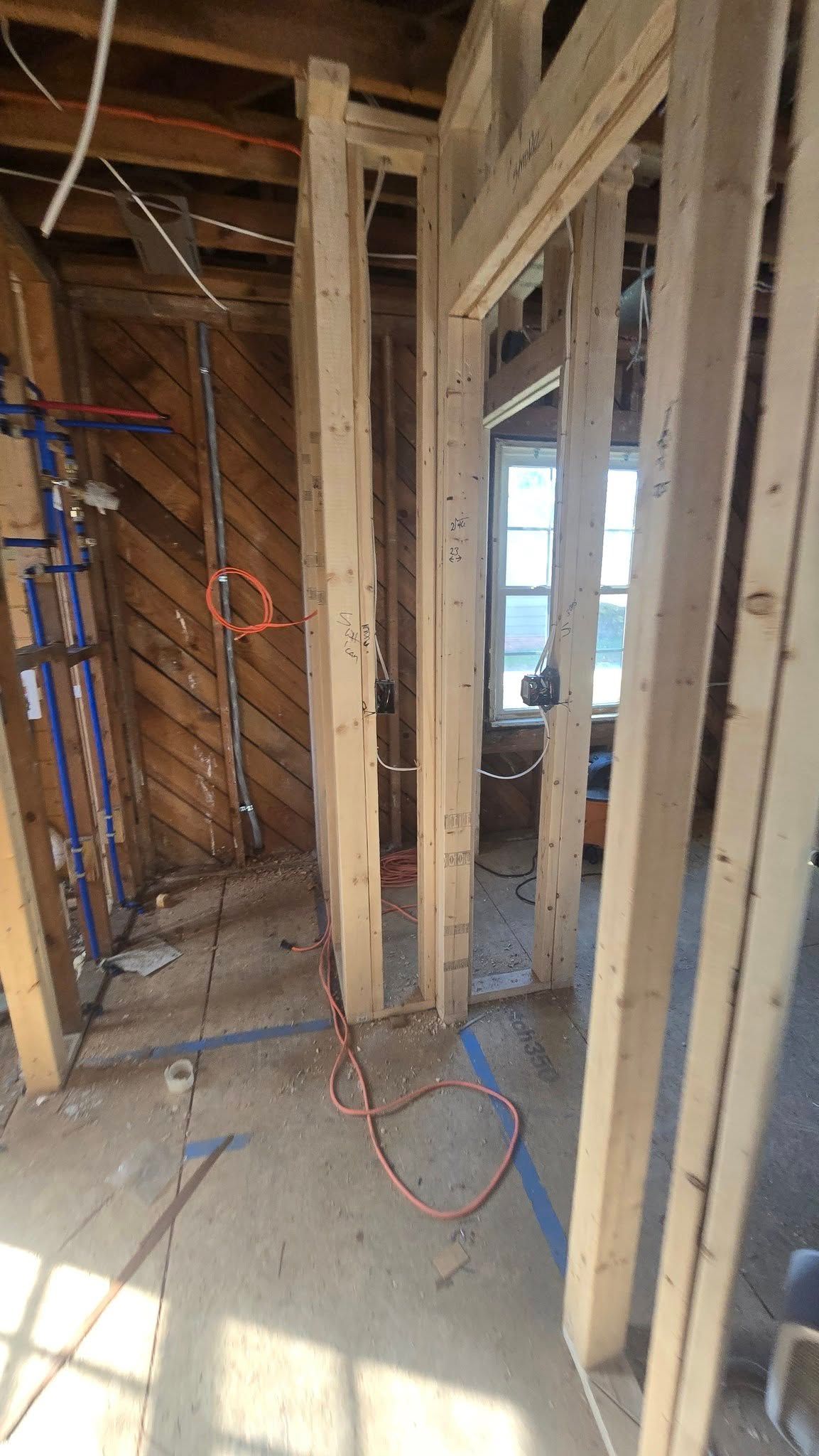 Interior wall framing with exposed plumbing and wiring; window in the background.