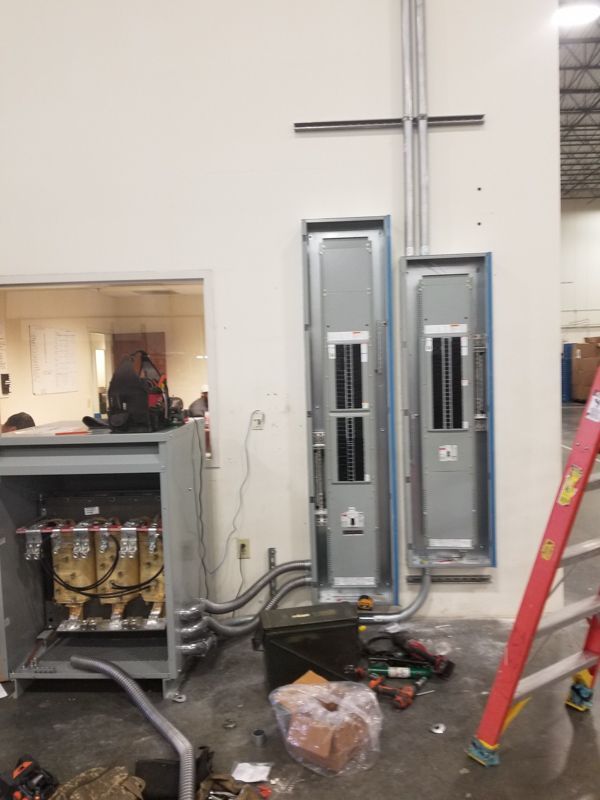 Electrical panels and exposed wiring in a utility room during construction, with tools and debris on the floor