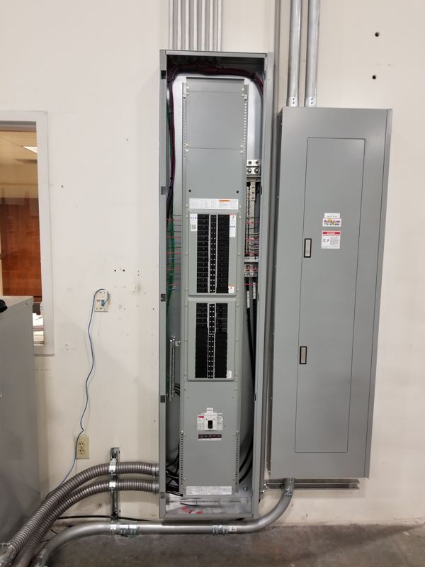 Open electrical panel and breaker cabinet in a utility room, with conduit running along the wall.