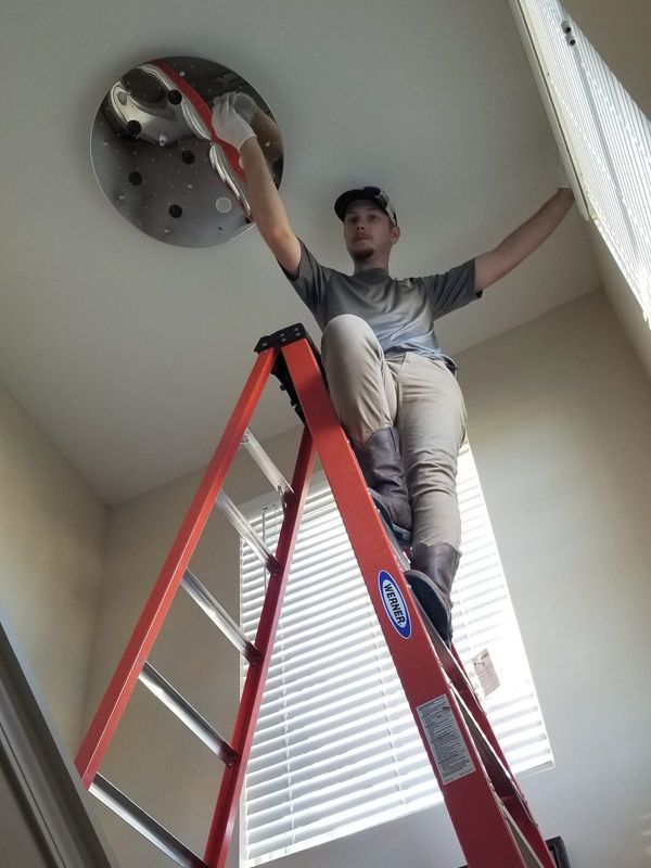 Person standing on a red ladder reaching up to work on a ceiling fixture in a room with blinds.