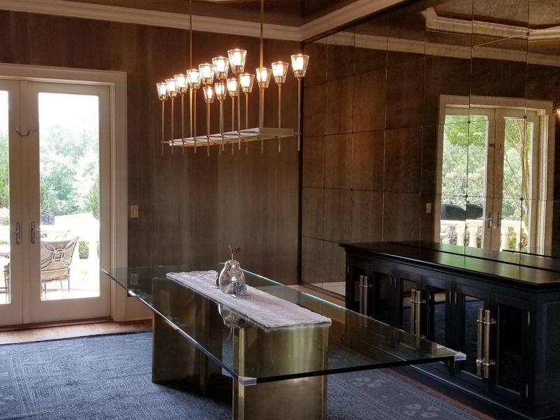 Modern dining room with a long glass table, chandelier, wood-paneled walls, and French doors to outside