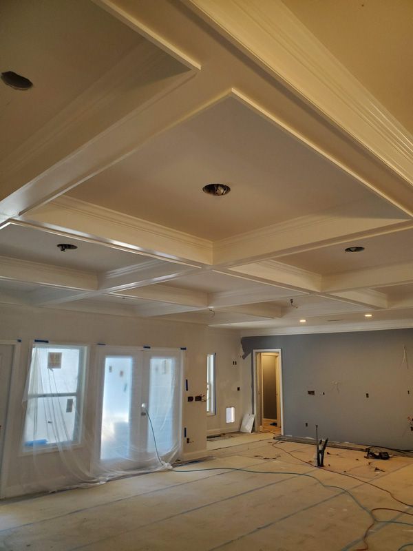 Empty room under renovation with recessed ceiling panels, covered floors, and taped windows and doorway.