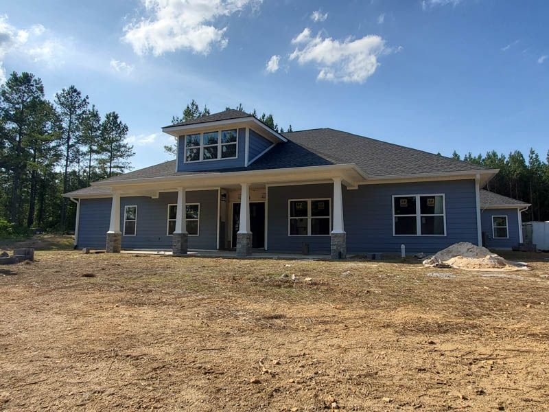 New blue house with front porch and dormer windows on a bare dirt lot under a clear sky