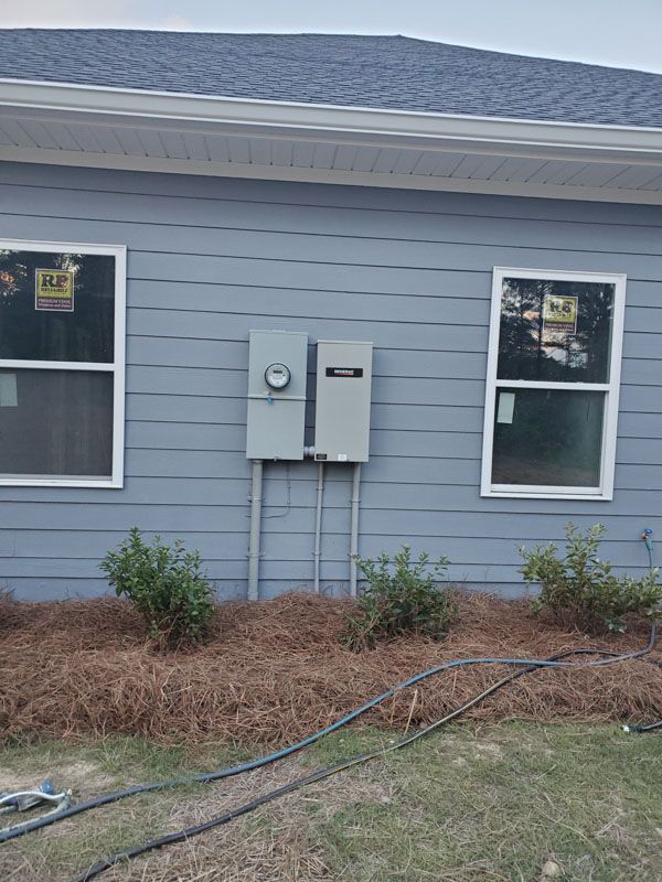 Gray house exterior with two windows, utility meters, and hoses in the grass near shrubs