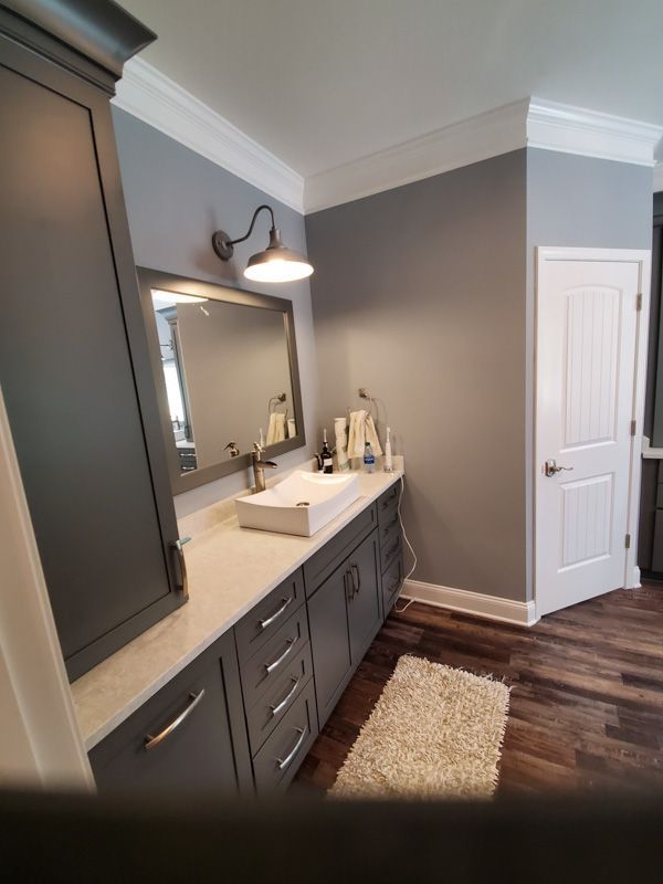 Gray bathroom vanity with large mirror, sink, wall sconce, white door, and dark wood floor with rug