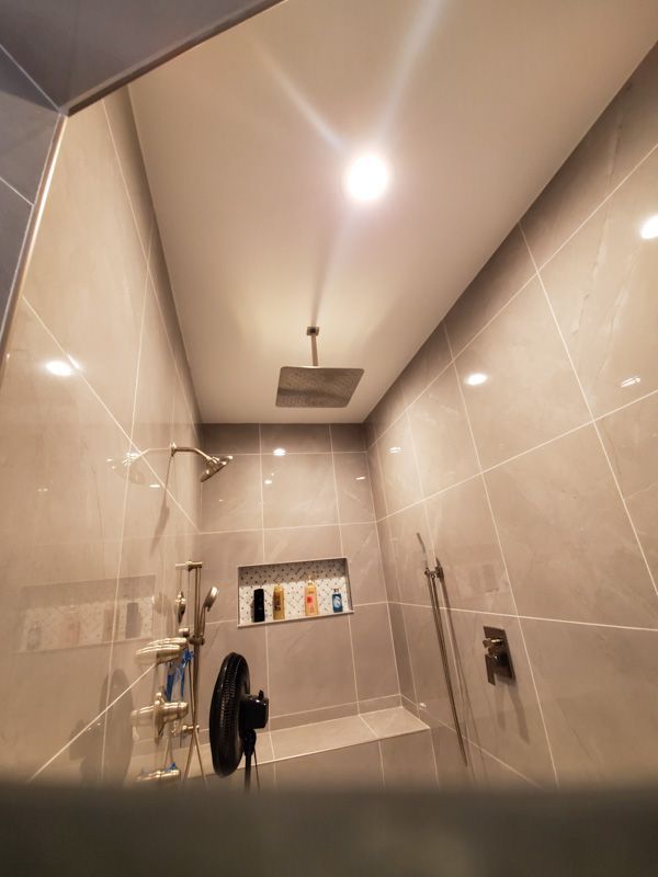 Narrow tiled shower stall with ceiling light, wall-mounted showerhead, and cleaning supplies on shelves.