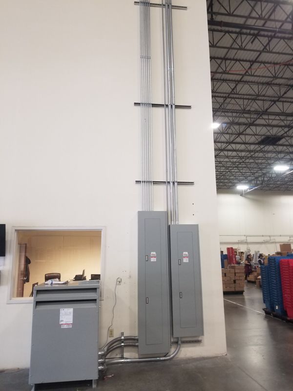 Industrial electrical panels and conduit mounted on a warehouse wall, with a small gray cabinet nearby.