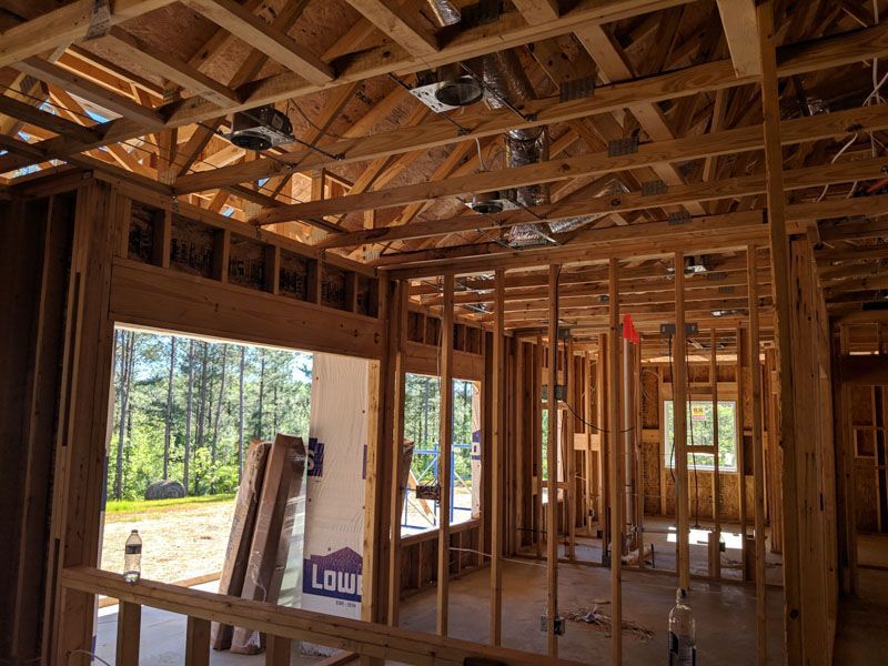 Unfinished wooden house interior with exposed framing, large windows, and sunlight streaming in.