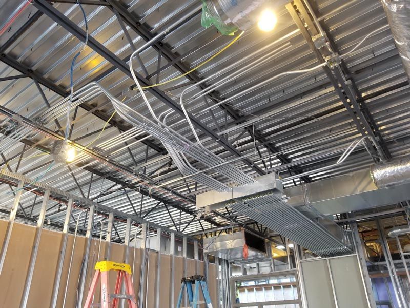 Indoor construction site ceiling with exposed metal framing, ducts, wiring, and hanging lights; ladder below