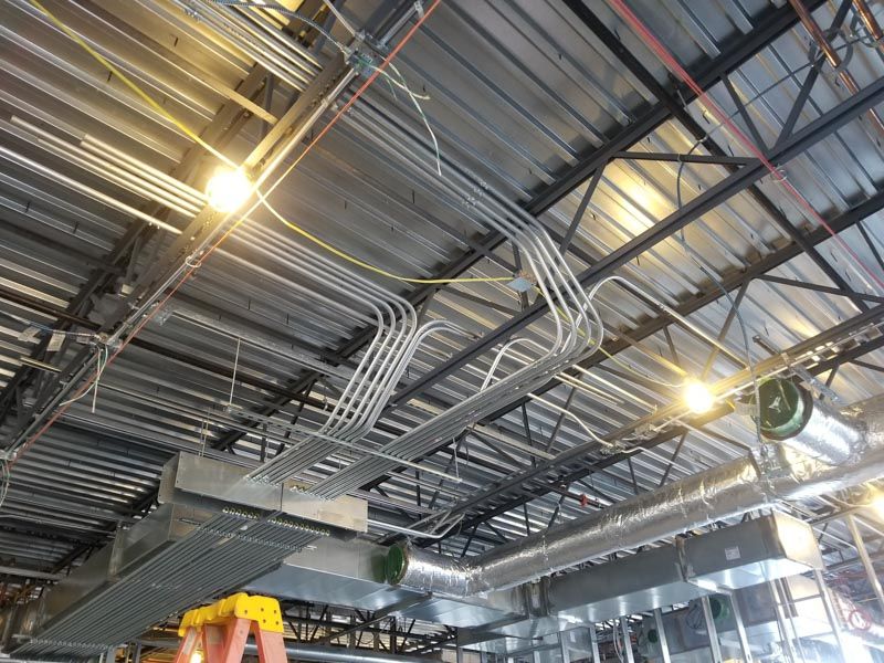 Industrial ceiling with exposed metal beams, ductwork, pipes, and bright hanging lights in a warehouse.