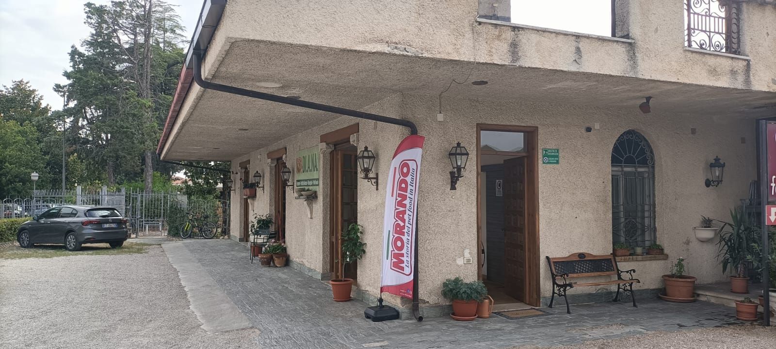 Un piccolo edificio a bordo strada con un distributore automatico rosso, piante in vaso e un'auto parcheggiata vicino a un piazzale ghiaioso.
