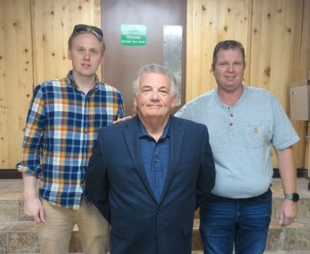 Three men standing indoors in front of a wood-paneled wall, wearing casual shirts and jackets.
