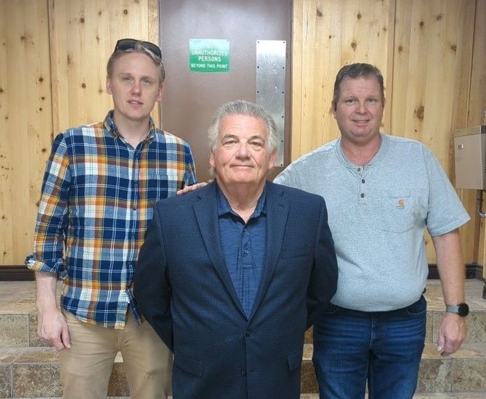 Three men standing indoors in front of a wood-paneled wall, wearing casual shirts and jackets.