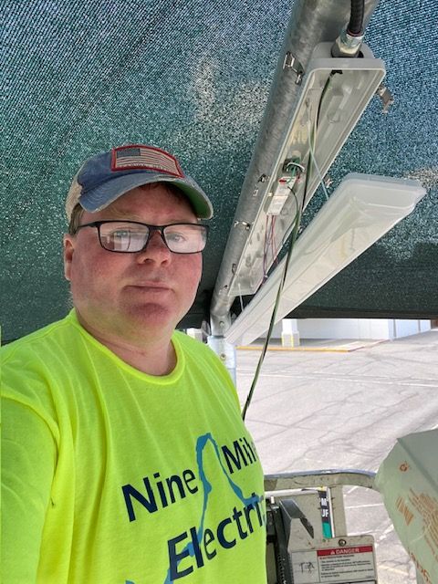 A person in a bright yellow work shirt and cap working on an outdoor light fixture under a green canopy.
