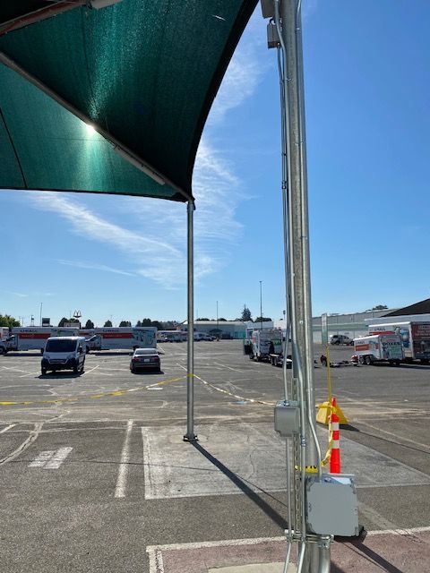 A view of a parking lot under a green shade canopy, featuring a metal pole with electrical boxes and several vehicles.