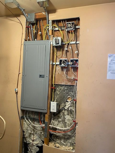 An electrical panel mounted on a wall with exposed wiring, junction boxes, and spray foam insulation in an open stud bay.