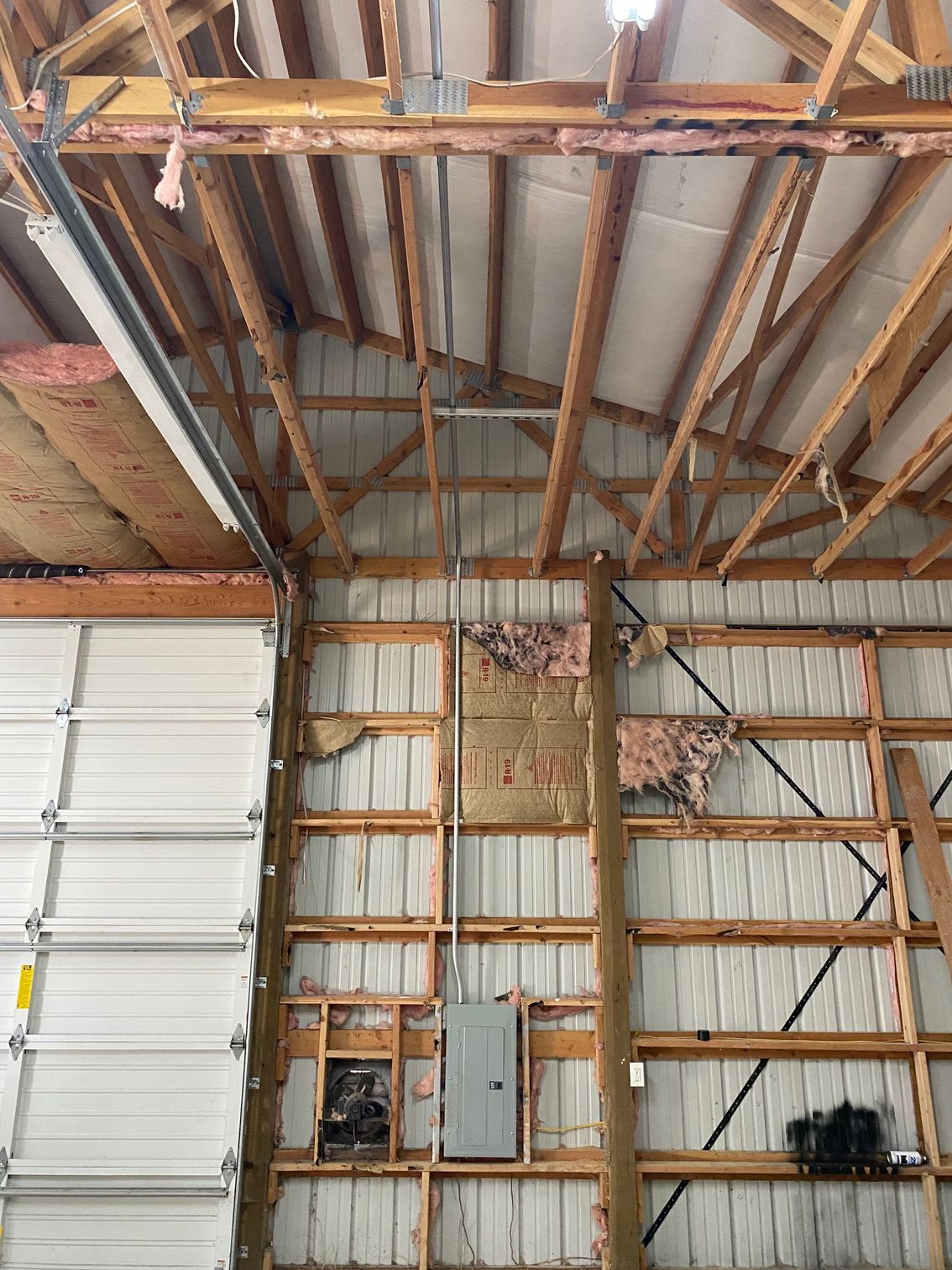 Interior view of a workshop wall with exposed wooden framing, insulation, a garage door, and an electrical panel.