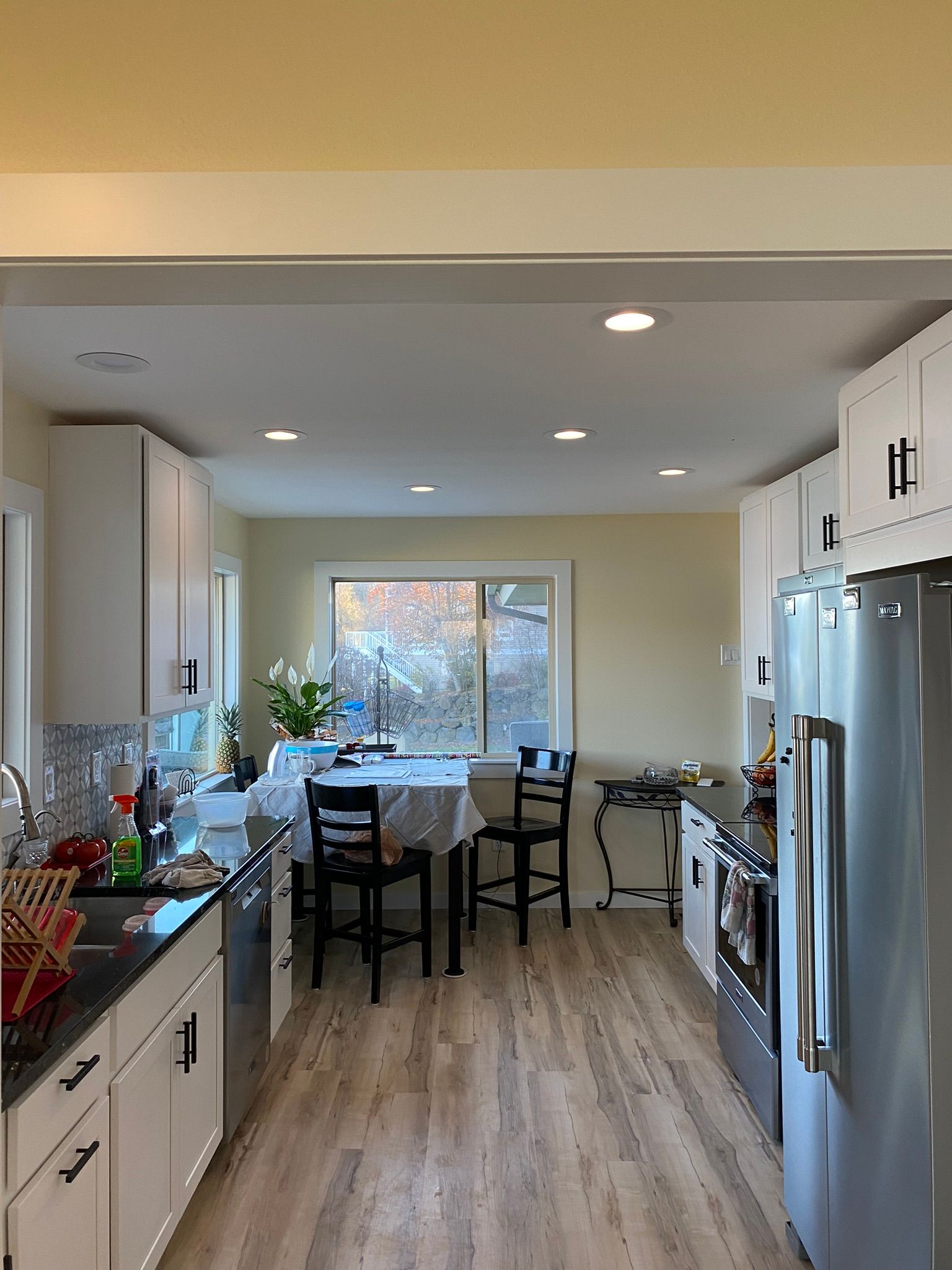 A kitchen with white cabinets, stainless steel appliances, and a small dining table by a window under recessed lighting.