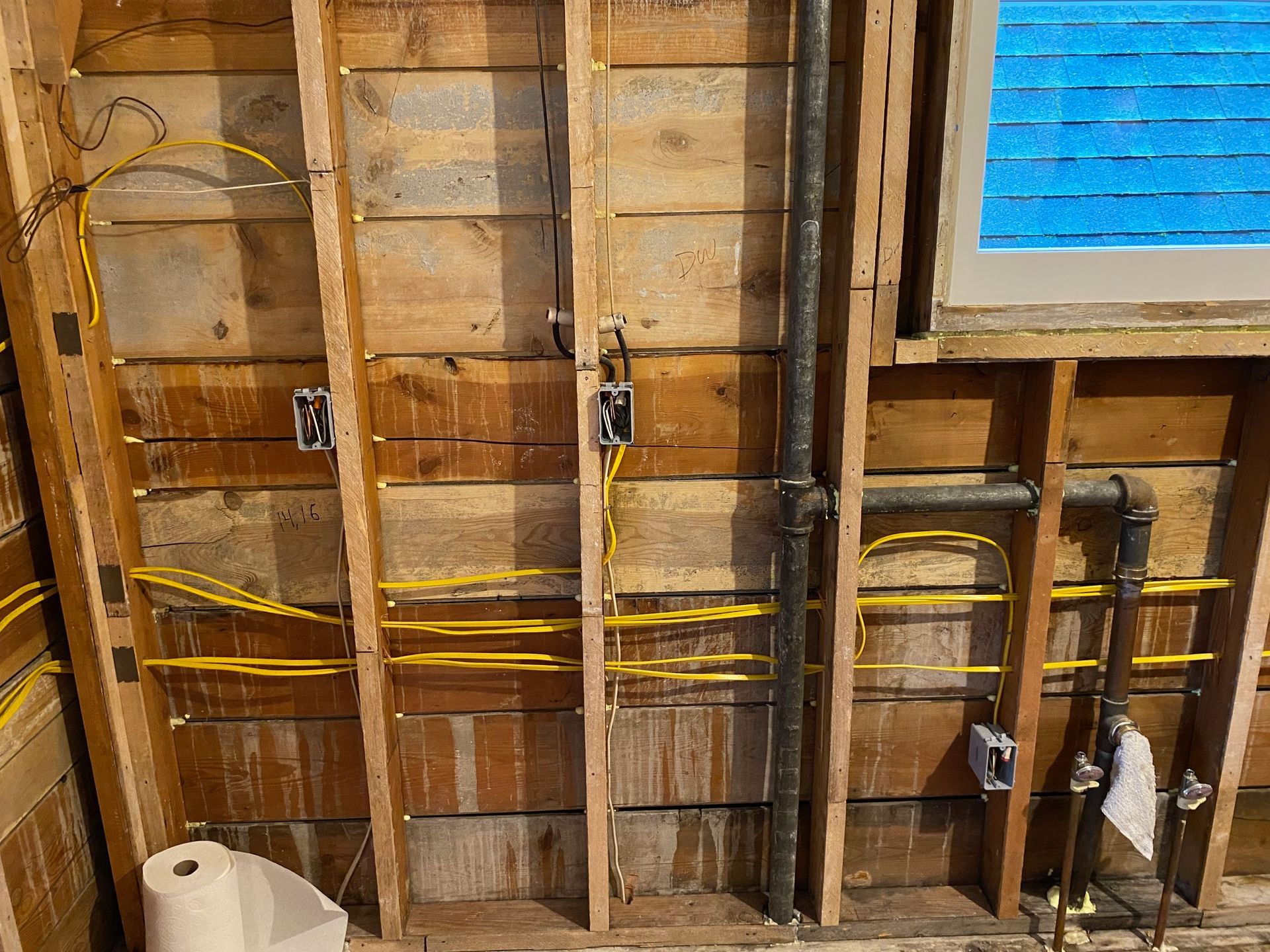 An interior wall under construction showing wooden studs, electrical wiring, plumbing pipes, and an adjacent window.