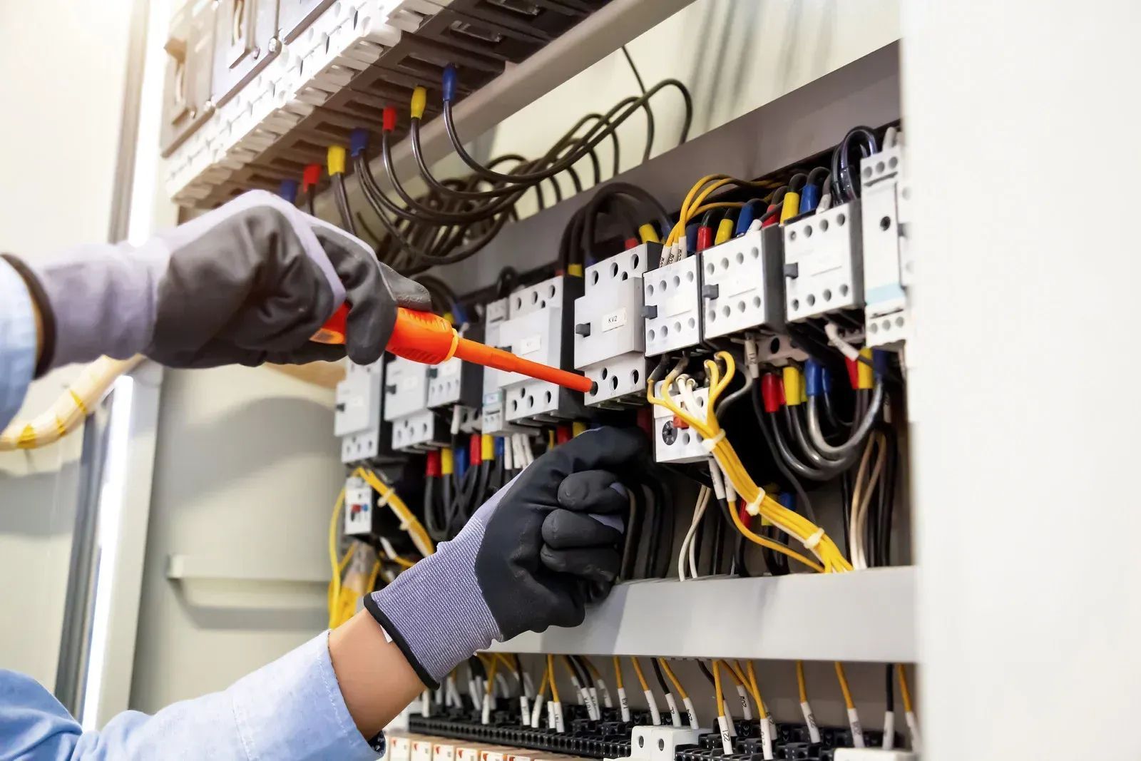 A person in work gloves uses a voltage tester on wires inside an electrical control panel.