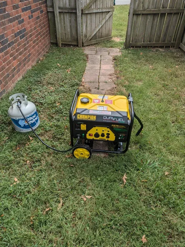A yellow Champion dual-fuel portable generator connected to a propane tank, sitting on the grass near a brick wall.