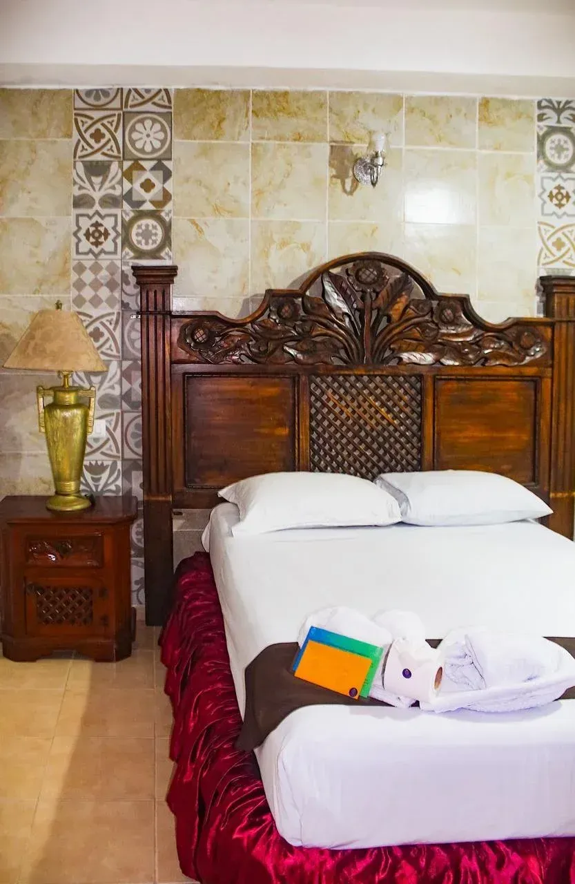 Bedroom with ornate wooden headboard, bedside table, white bedding, and patterned wall tiles.