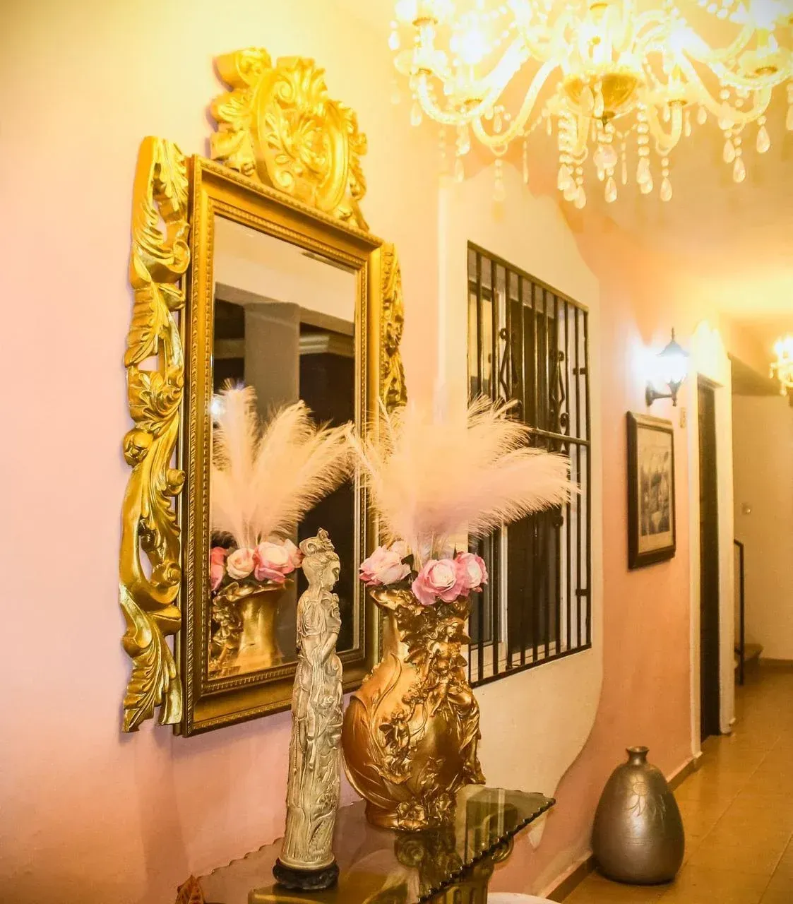 Ornate gold mirror, vases, and sculpture on a table in a hallway with pink walls.
