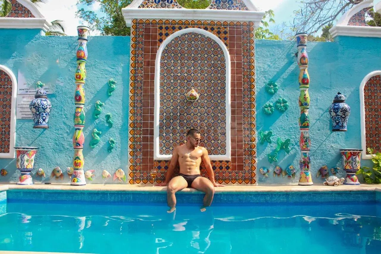 Man sitting on the edge of a turquoise pool, with ornate wall and mosaic decorations in the background.
