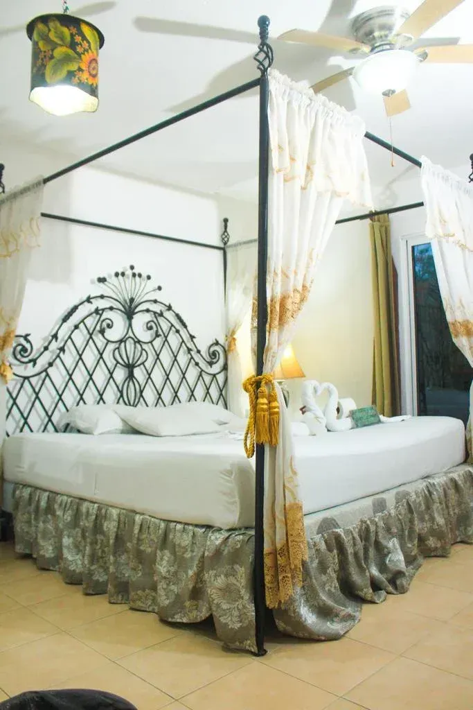 Four-poster bed in a room; ornate black headboard, white linens, floral skirt, gauzy curtains, ceiling fan, and pendant lamp.