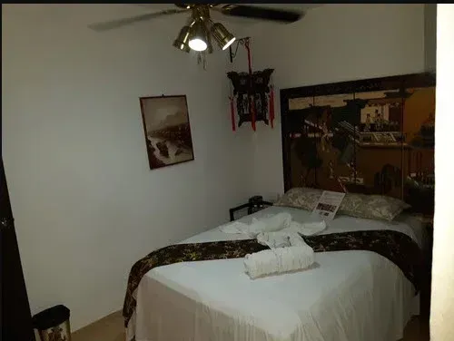 Dormitorio con ropa de cama blanca, cabecera marrón, obras de arte, lámpara decorativa y ventilador de techo.