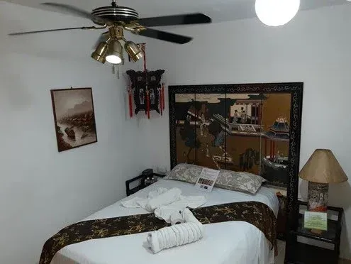 Dormitorio con cama, cabecero oscuro, ventilador de techo y decoración de temática asiática.