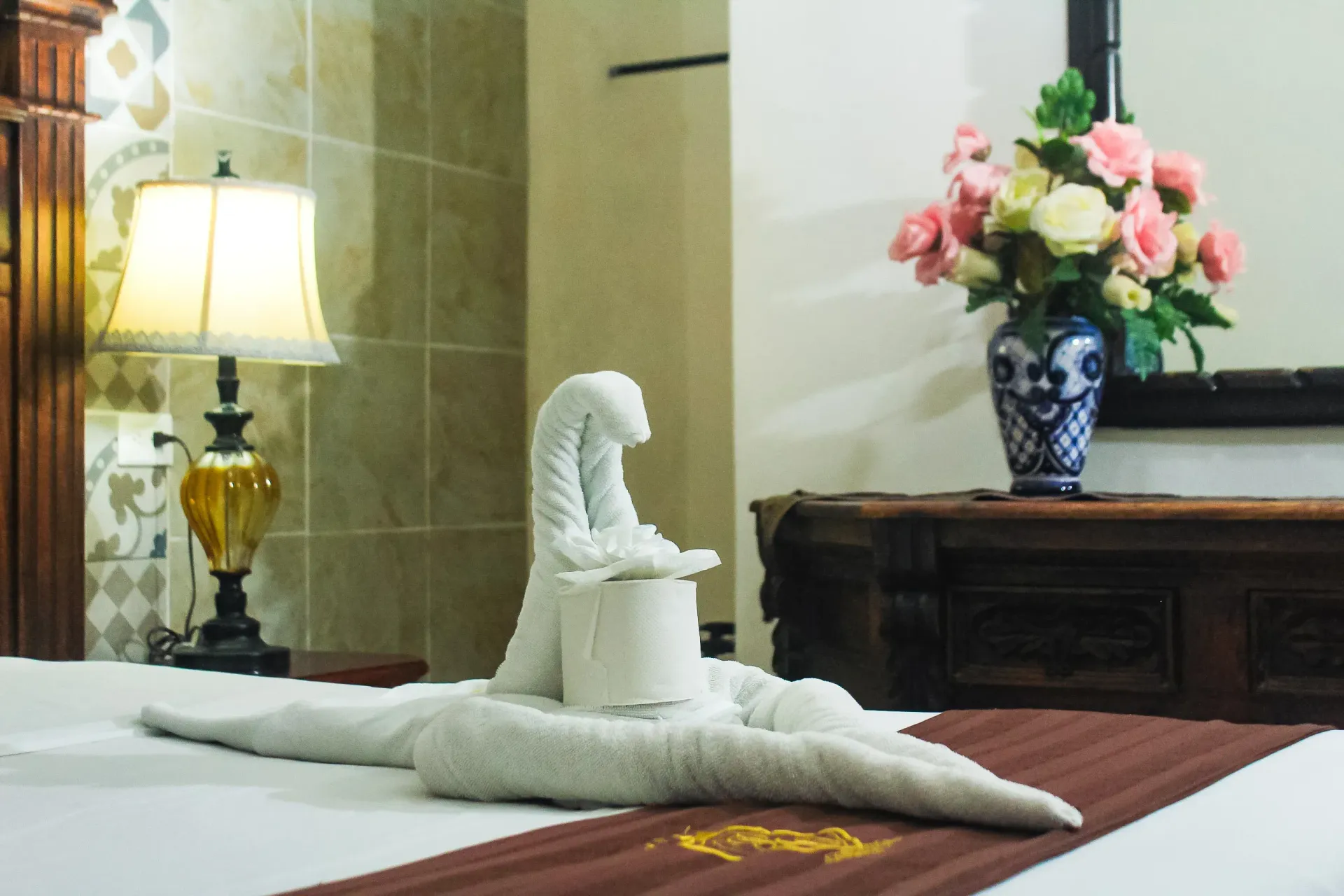 Cama de habitación de hotel con toalla de cisne, lámpara, flores en jarrón y muebles de madera oscura.