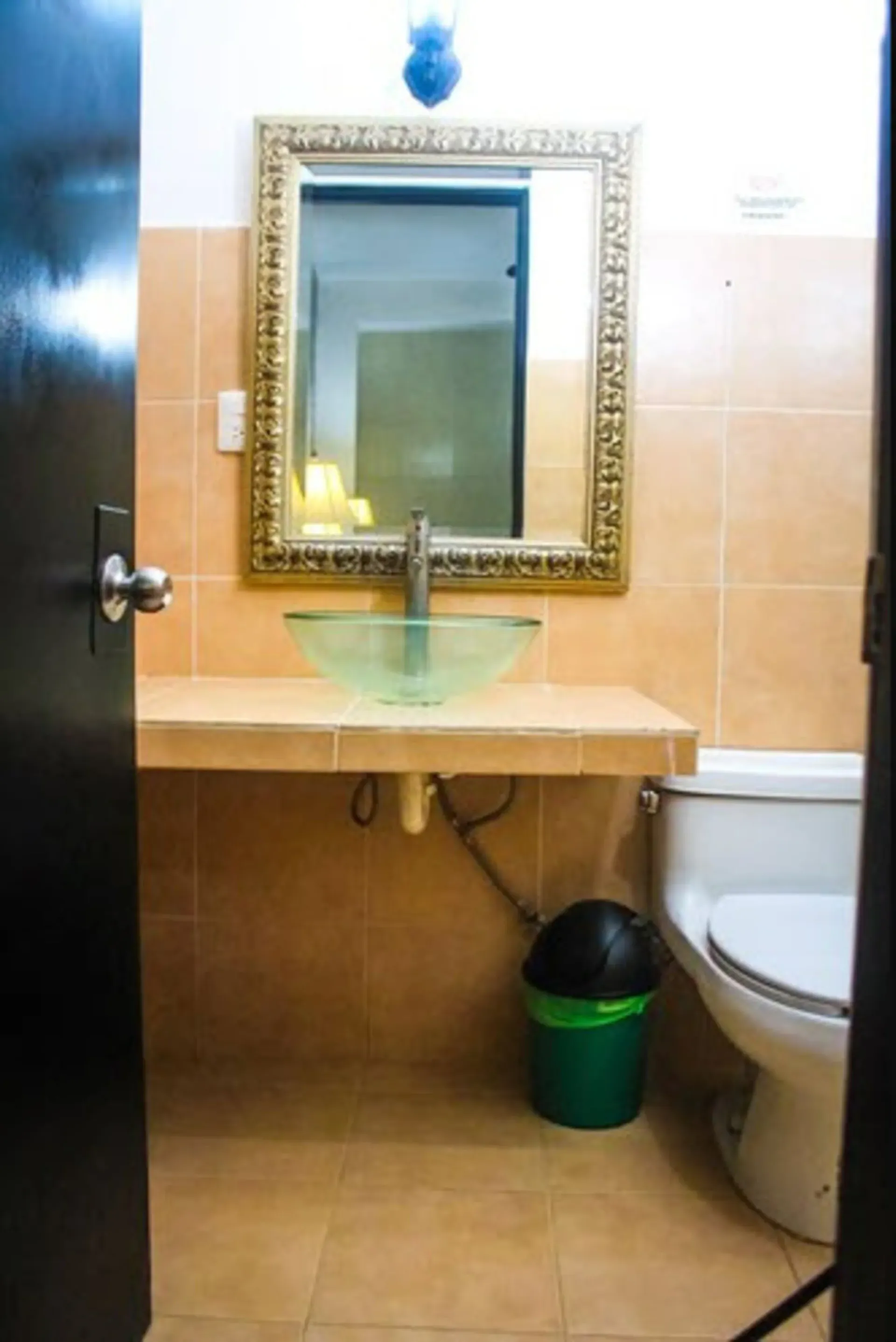 Baño con espejo rectangular con marco dorado, lavabo de cristal e inodoro. Las paredes son de color melocotón.