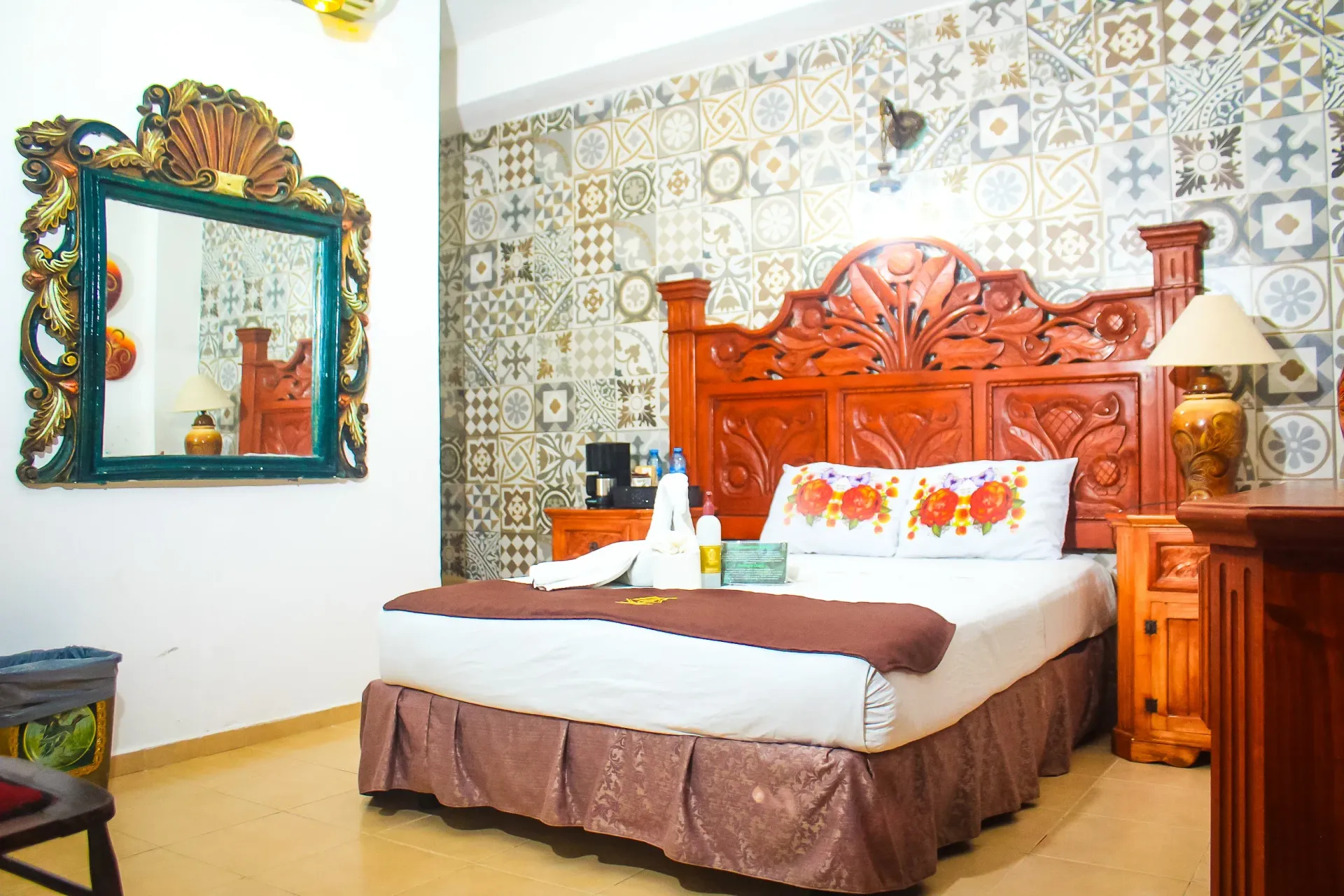 Dormitorio con cabecero de madera ornamentado, espejo grande, azulejos de pared estampados y colcha marrón.