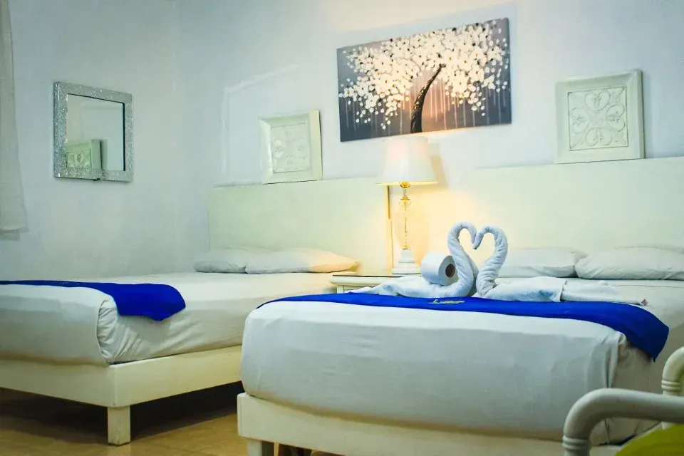 Habitación de hotel blanca con dos camas, detalles azules y obras de arte.