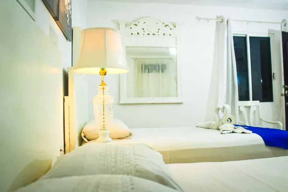 Dormitorio blanco con dos camas individuales, lámpara, espejo y ventana con cortinas.