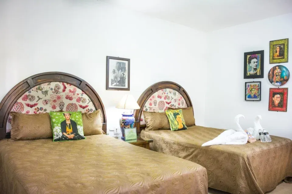 Dos camas individuales en una habitación luminosa con cabeceras florales, almohada de Frida Kahlo y arte en la pared.
