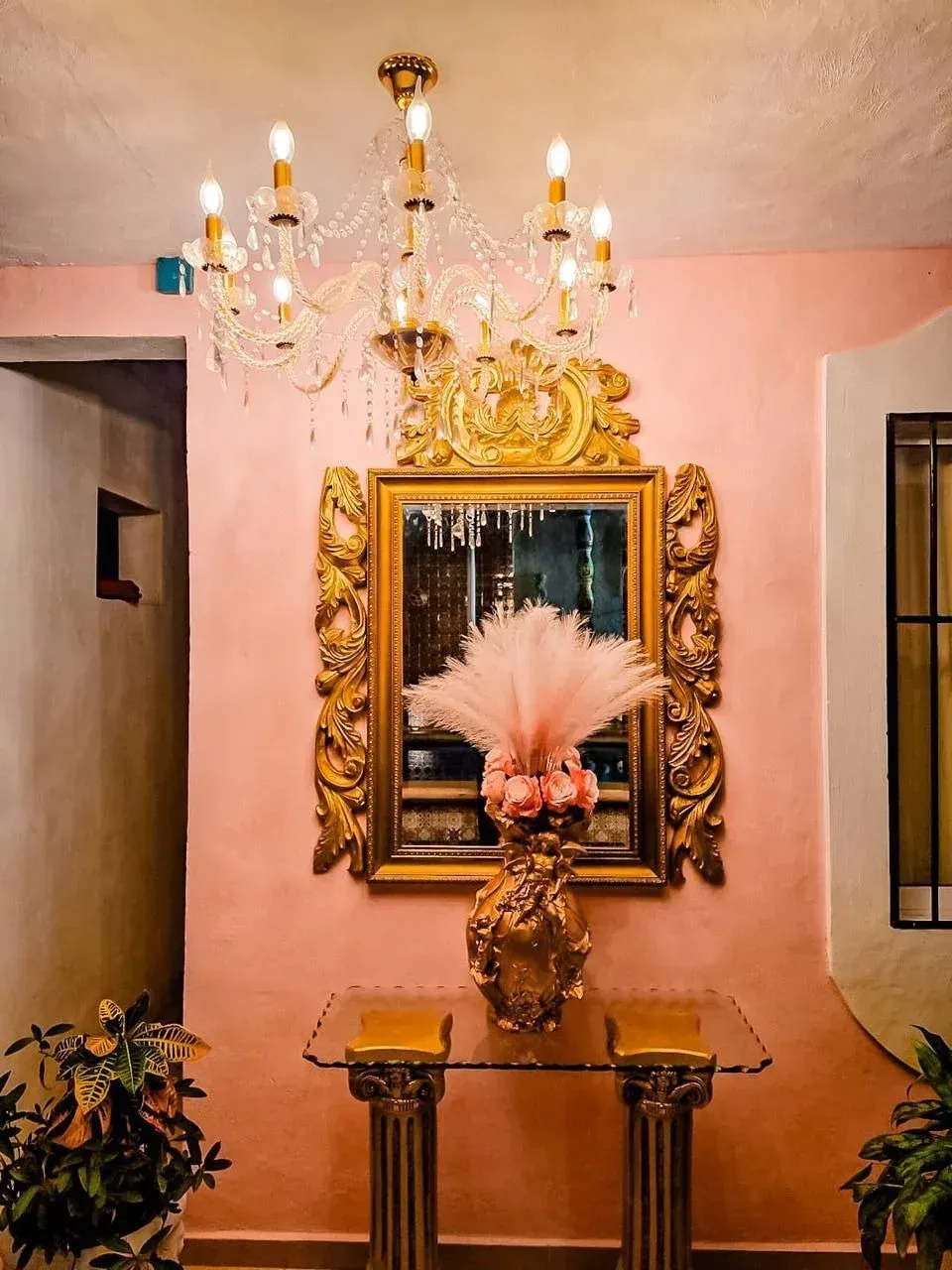 Ornate mirror and chandelier on pink wall, glass table with floral arrangement.