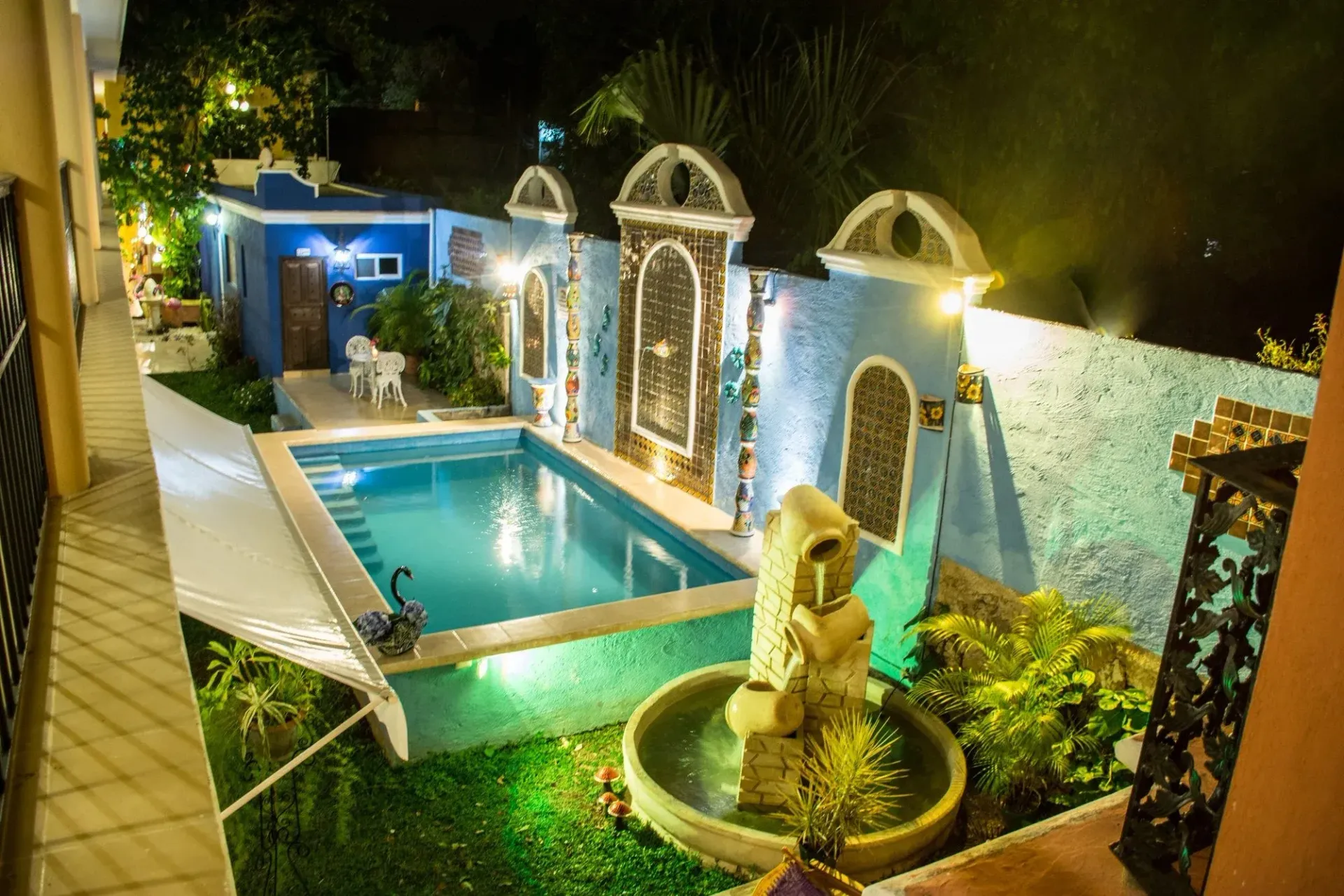 Nighttime view of a turquoise pool, fountain, and ornate blue wall with archways; lush greenery and lights.