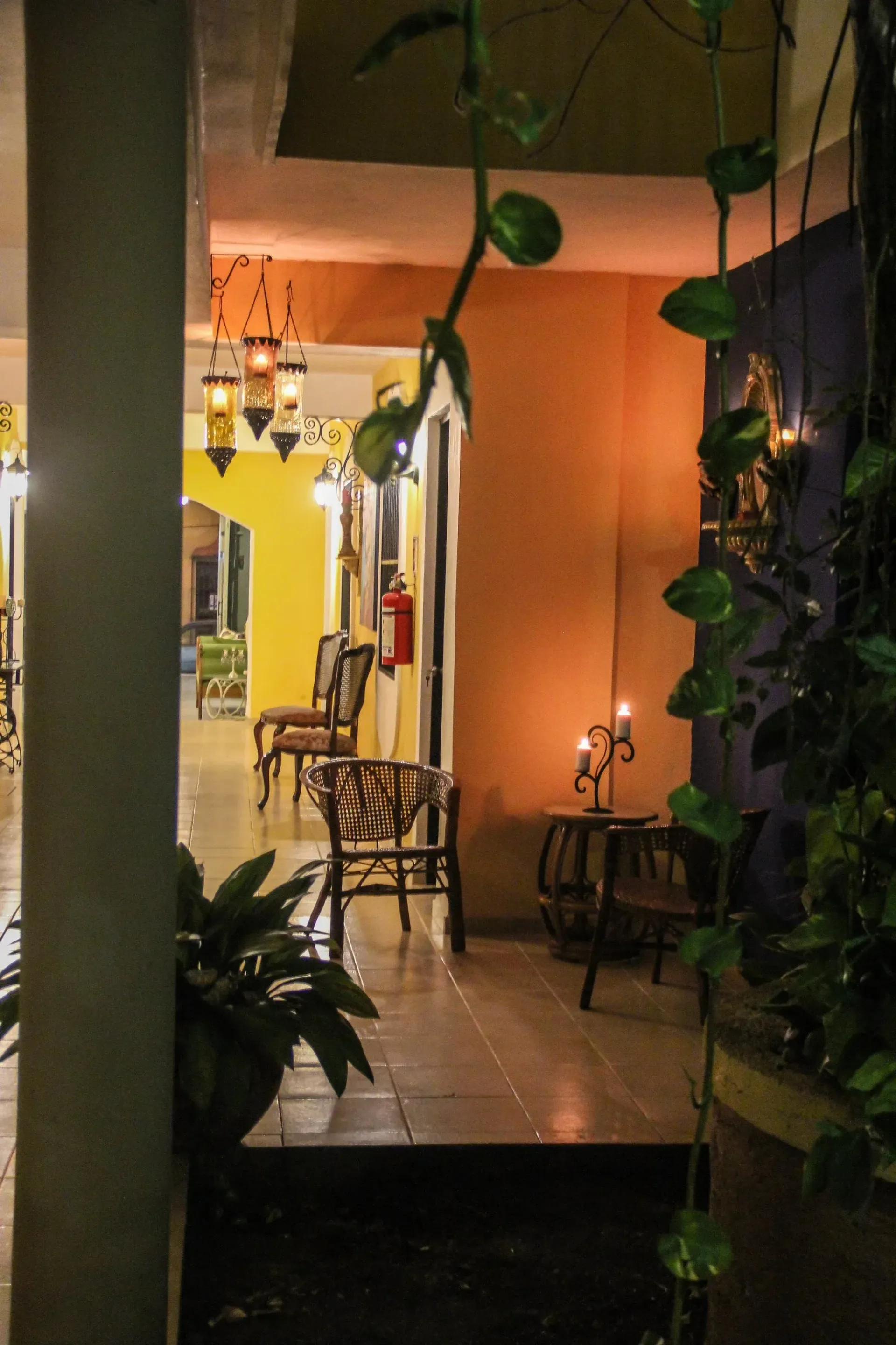 Warmly lit hallway, chairs, and potted plants with vines. Candles on a small table.