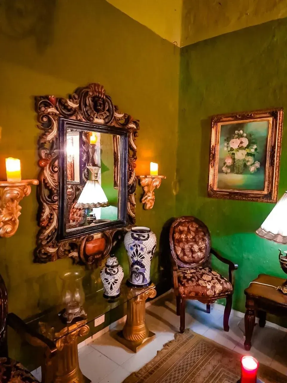 Ornate mirror, vases, chair, and art in a dimly lit room with green walls.