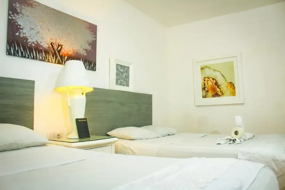 Two beds in a white-walled room with art, a lamp, and a nightstand.