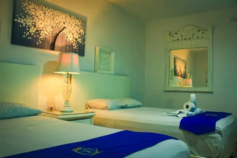 Twin beds in a white-walled room with a lamp, artwork, and mirror. Blue accents.