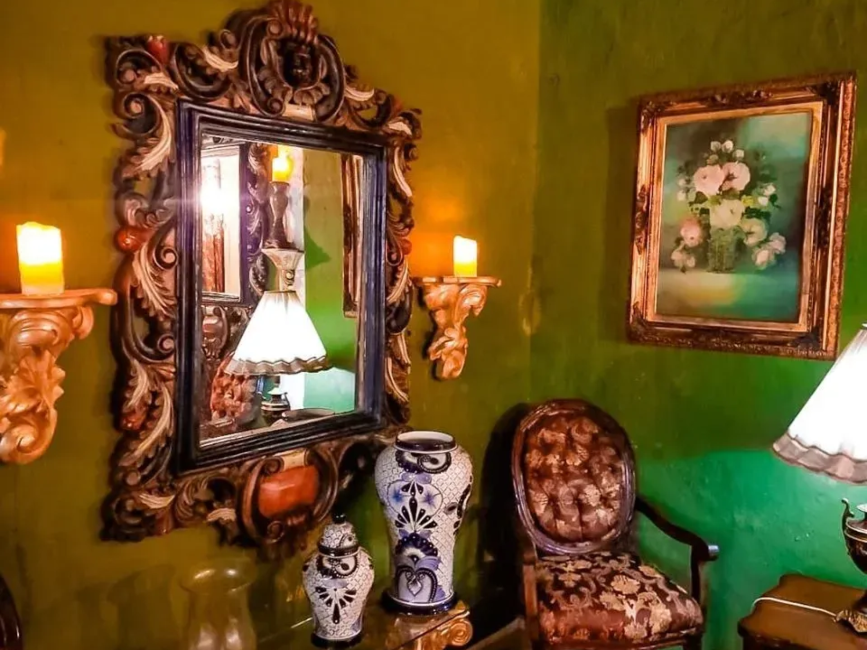 Ornate mirror, candles, paintings, and vases in a room with green walls, creating a vintage atmosphere.