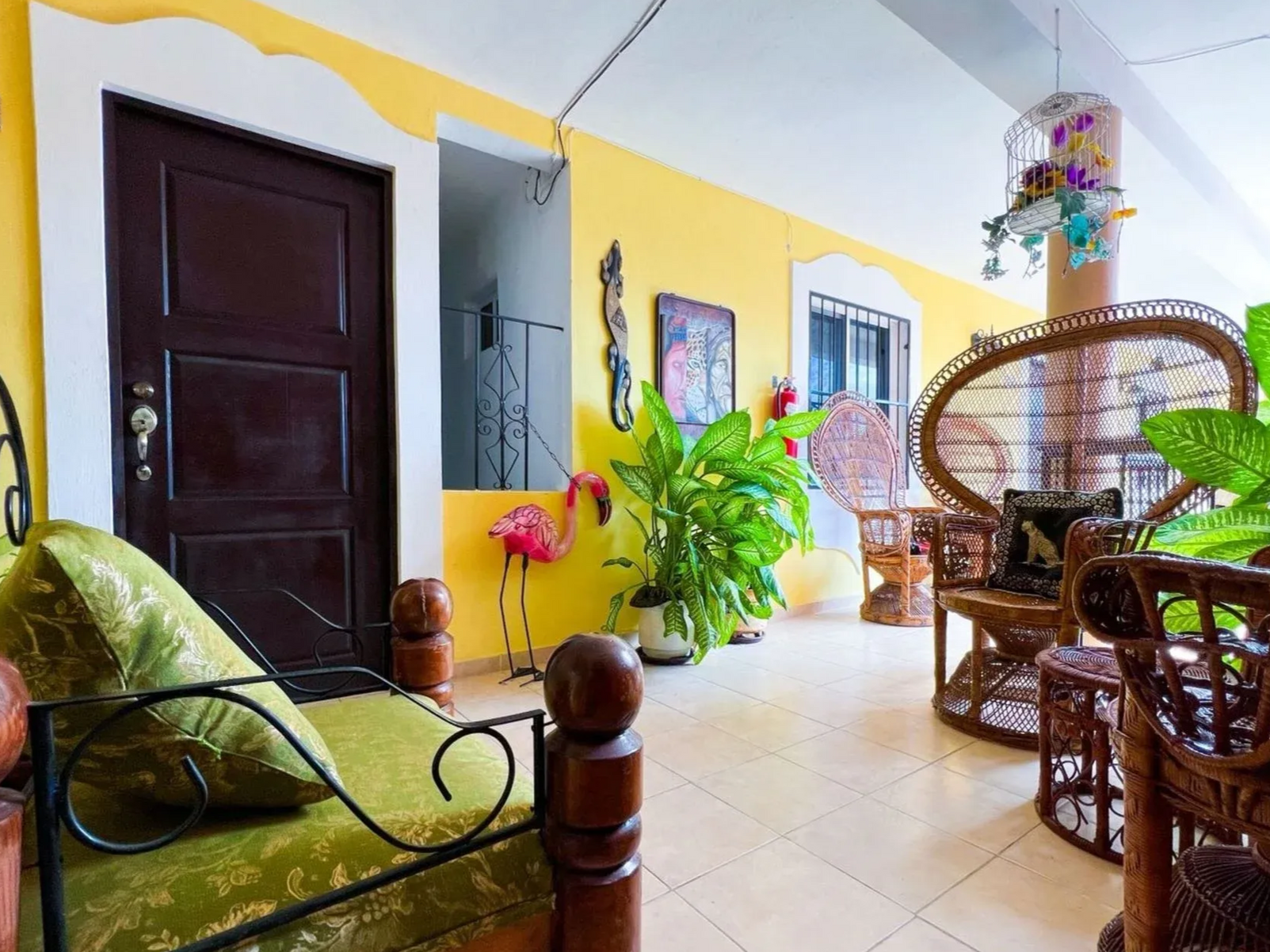 A vibrant, yellow-walled veranda with wicker furniture, plants, and a dark brown door.